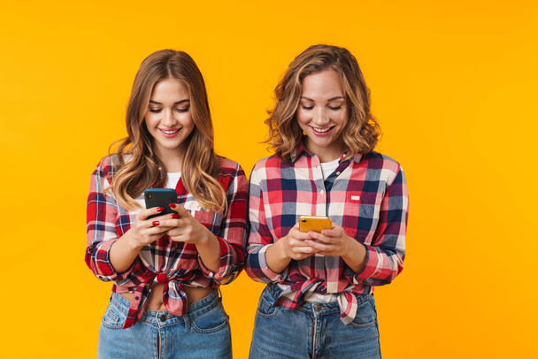 Image of two young beautiful girls wearing plaid shirts smiling and using cellphones isolated over yellow background - Foto, afbeelding