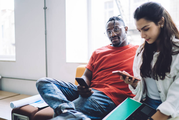Confident handsome black man sitting on couch with diverse female colleague using mobile phone and looking at camera in office hall - Foto, Imagen
