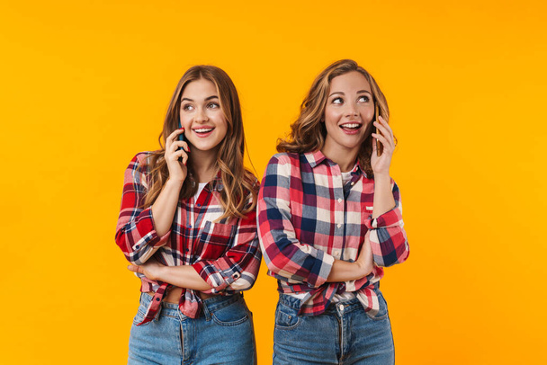 Image of two young beautiful girls wearing plaid shirts smiling and using cellphones isolated over yellow background - Foto, immagini