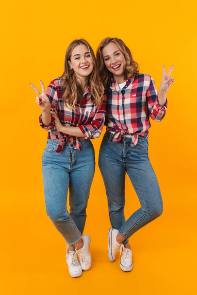 Full length image of two young beautiful girls wearing plaid shirts smiling and gesturing peace sign isolated over yellow background - Foto, imagen