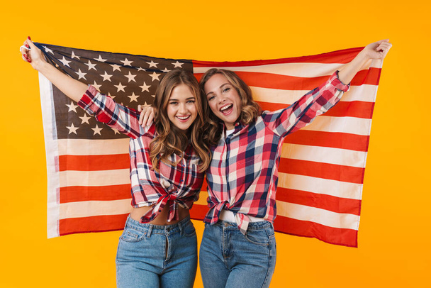 Image of two young beautiful girls wearing plaid shirts smiling and american flag together isolated over yellow background - Photo, Image