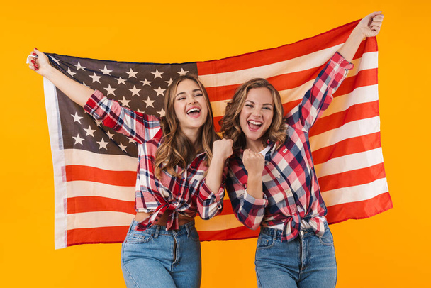 Image of two young beautiful girls wearing plaid shirts smiling and american flag together isolated over yellow background - Фото, зображення