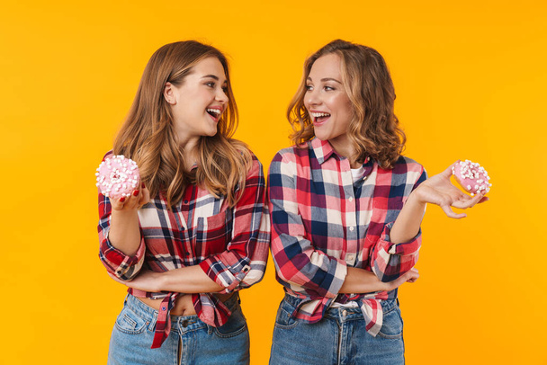 Image of two young beautiful girls wearing plaid shirts having fun and eating donuts isolated over yellow background - Foto, immagini