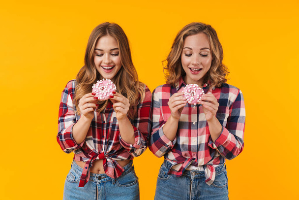 Image of two young beautiful girls wearing plaid shirts having fun and eating donuts isolated over yellow background - Foto, imagen