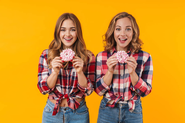 Image of two young beautiful girls wearing plaid shirts having fun and eating donuts isolated over yellow background - Valokuva, kuva