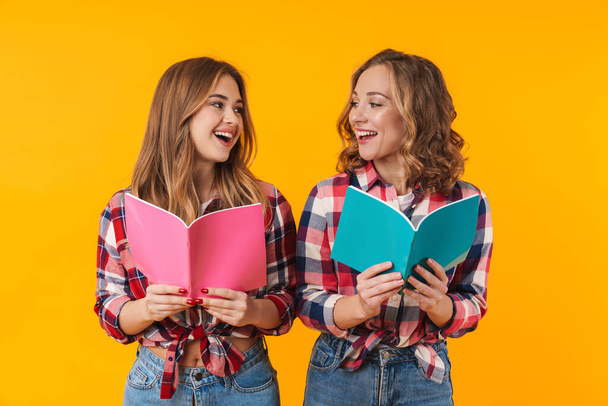 Image of two young beautiful girls wearing plaid shirts smiling and holding diary books isolated over yellow background - Fotó, kép