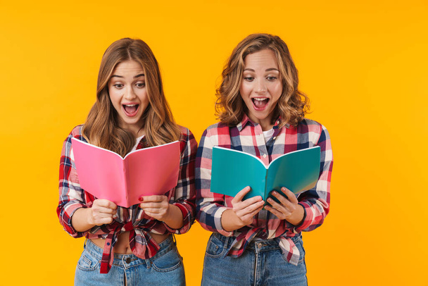 Image of two young beautiful girls wearing plaid shirts smiling and holding diary books isolated over yellow background - Foto, Imagen