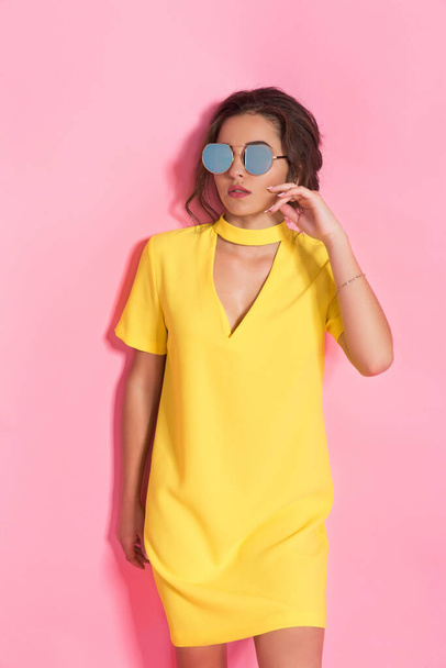 Beautiful girl wearing yellow dress and sunglasses posing on pink background in studio - Photo, image