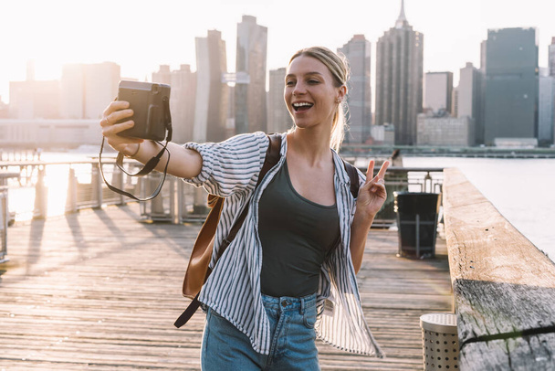 Happy woman traveller with retro technology having fun during vacations in United States showing v-sign while clicking selfie picture with Manhattan district on background, recreating journey - Photo, Image