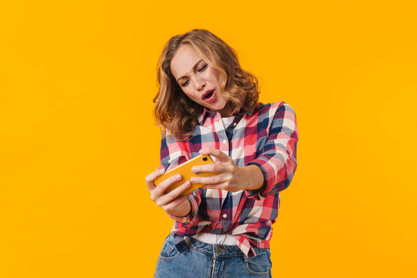 Image of young beautiful woman wearing plaid shirt smiling and holding cellphone isolated over yellow background - Photo, Image