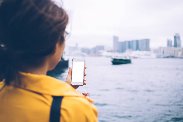 Back view of unrecognizable woman wearing yellow shirt holding white cellphone with empty screen standing on bank of city bay in Hong Kong on nasty day - Фото, изображение