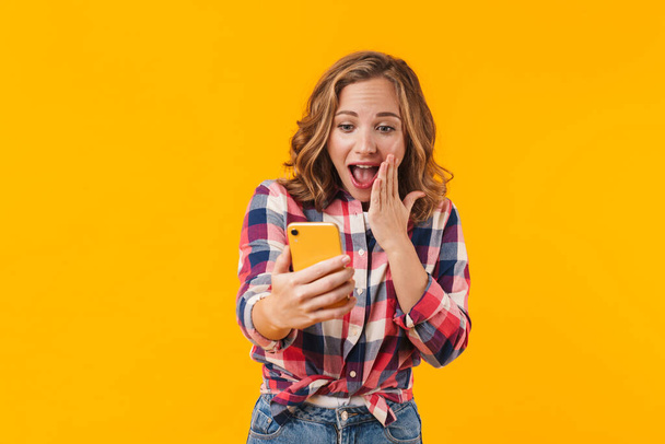 Image of young beautiful woman wearing plaid shirt rejoicing and holding cellphone isolated over yellow background - Fotoğraf, Görsel