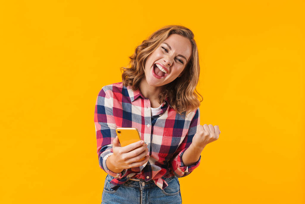 Image of young beautiful woman wearing plaid shirt rejoicing and holding cellphone isolated over yellow background - Fotografie, Obrázek