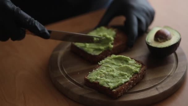 Close-up of Woman in black glowes puts guacamole or avocado spread on top of rye bread toast on wooden board at home. Vegan breakfast - Footage, Video
