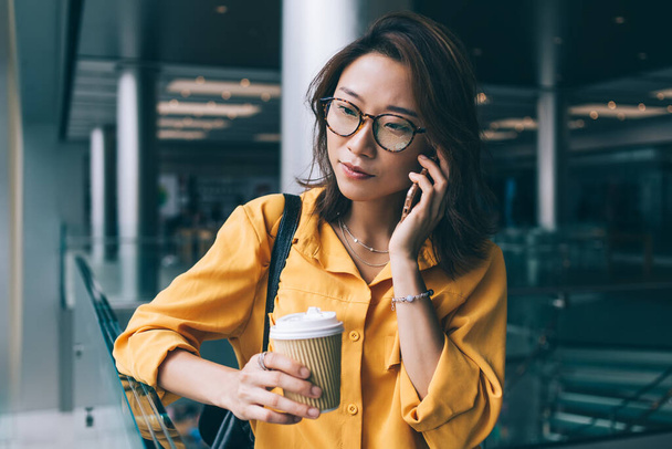 Beautiful thoughtful slender Asian woman in glasses with carton cup of coffee talking on phone looking away in mall in Hong Kong - Foto, immagini