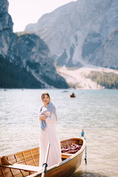 花嫁はイタリアのラーゴ・ディ・ブライス(Lago di Braies)の木製ボートに立っています。ヨーロッパでの結婚式、ブライス湖で。白いウェディングドレスを着た若い女の子が肩を青いセーターで覆い. - 写真・画像