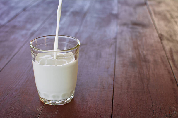 Ein Strom frischer Milch wird in einen Glasbecher gegossen. Ein Glas mit Milch steht auf einem alten Holztisch. Ernährungskonzepte für Naturprodukte und Gesundheit. - Foto, Bild