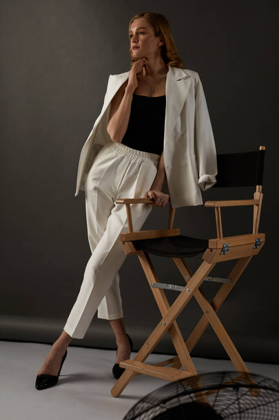 Female Fashion Portrait. Beautiful Woman In White Suit Stands Near The Wooden Director's Chair. Elegant High Fashion Clothes. - Фото, изображение