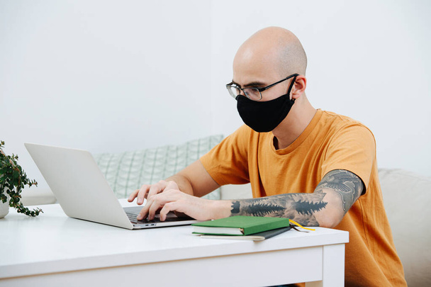 Bold freelancer in glasses and handmade black mask working on his laptop behind the table at home during covid-19 isolation. - Photo, image