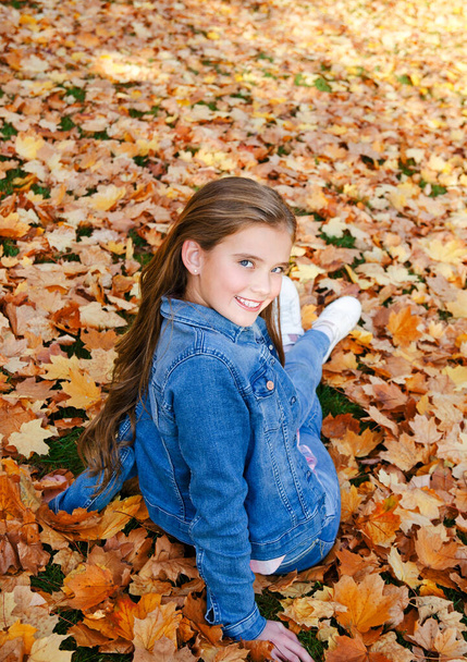 Осенний портрет очаровательной улыбающейся маленькой девочки-подростка, сидящей в листьях в парке на открытом воздухе
 - Фото, изображение
