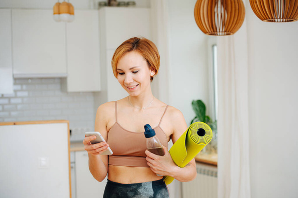 Happy woman texting after yoga practice. She's holding mat and water bottle. She's home in her kitchen. - Foto, imagen