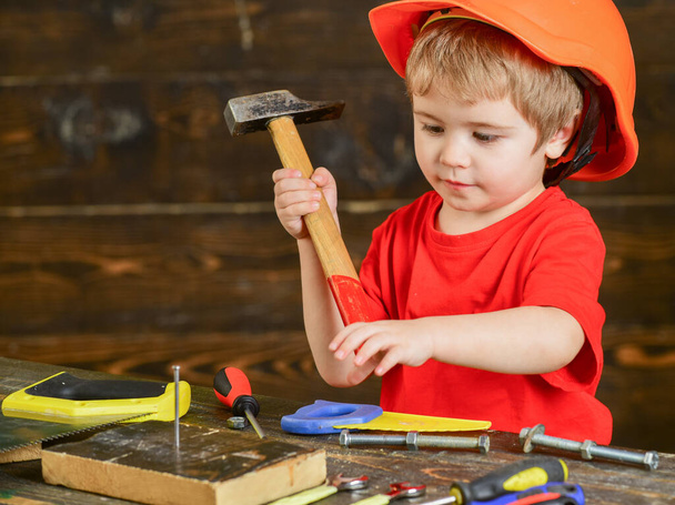 Little boy in orange helmet hammering nails into wooden board. Cute kid in workshop. Manual labor concept - 写真・画像