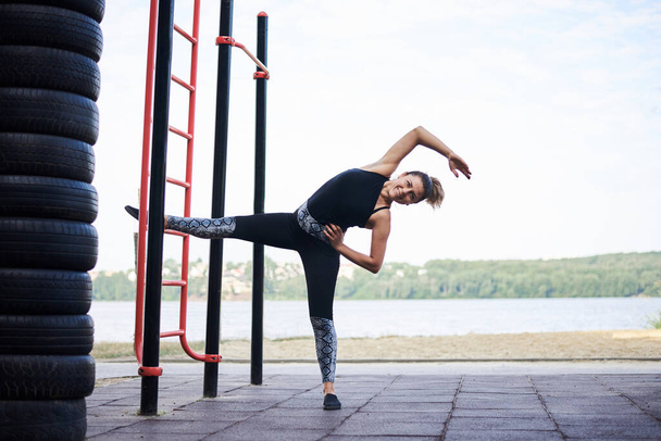 Young brunette woman, wearing black fitness overall, doing stretching exercises at swedish wall by city lake in summer. Fit sportswoman on outside training. Healthy energetic lifestyle concept. - Foto, Imagem
