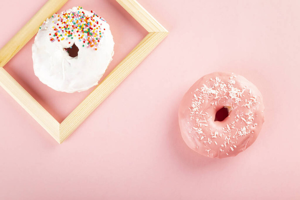Donut with white and pink icing and multicolored sugar pastry topping on pink background. One donut in wooden frame. Top view - Foto, Bild