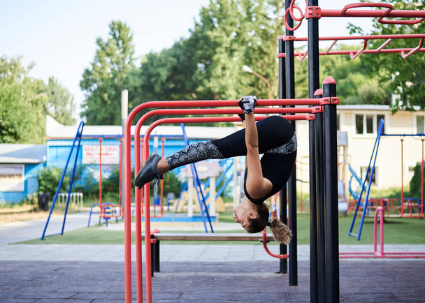 Young brunette woman, wearing black fitness overall, doing stretching exercises, hanging on horizontal bars on sports playground in summer. Fit sportswoman outside, training. Healthy lifestyle concept - Foto, afbeelding