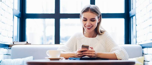 Positive hipster girl in casual apparel using cellphone gadget for chatting with virtual friends connecting to cafeteria wifi, happy cheerful blogger networking surf websites via smartphone device - Φωτογραφία, εικόνα