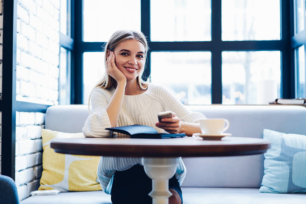 Portrait of happy hipster girl sitting at cafeteria table with smartphone device and smiling at camera during networking time, cheerful woman using 4g for phoning browsing website via cellular - Foto, afbeelding