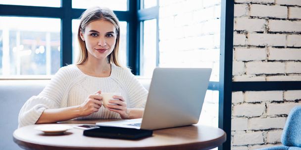 Happy woman with mug of caffeine beverage smiling at camera during time for using laptop for searching new friends in internet social networks, skilled female freelancer spending day for distance job - Photo, image