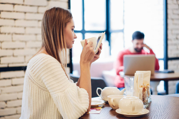 Side view of attractive female holding cup with hot tasty tea and writing email message via cellphone application connecting to wifi internet in coffee shop, millennial hipster girl checking media - Фото, изображение
