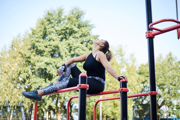 Young brunette woman, wearing black fitness overall, sitting on top of horizontal bars on sports playground in summer. Fit sportswoman outside, relaxing after training.Healthy lifestyle concept. - Zdjęcie, obraz