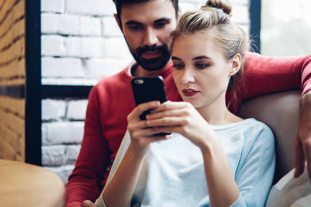 Millennial male and female embracing indoors and browsing websites via app enjoying blogging lifestyle, Caucasian couple connected to 4g on cellular for download media files via application - Fotó, kép