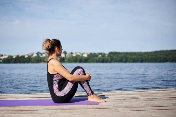 Young brunette woman with bare feet, wearing black and purple fitness outfit, stretching on violet yoga mat outside on wooden pier in summer. Fit girl, doing yoga poses by lake. Healthy lifestyle. - Photo, Image