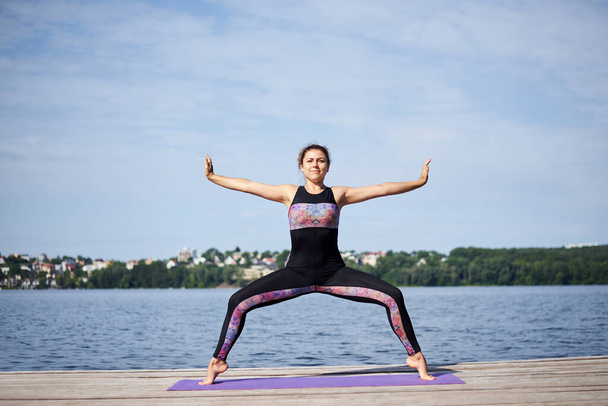 Young brunette woman with bare feet, wearing black and purple fitness outfit, stretching on violet yoga mat outside on wooden pier in summer. Fit girl, doing yoga poses by lake. Healthy lifestyle. - Foto, Bild