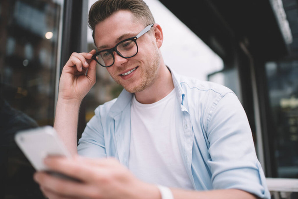 From below of cool millennial man in casual shirt and black glasses using smartphone and smiling confidently sitting at table on street - Fotoğraf, Görsel