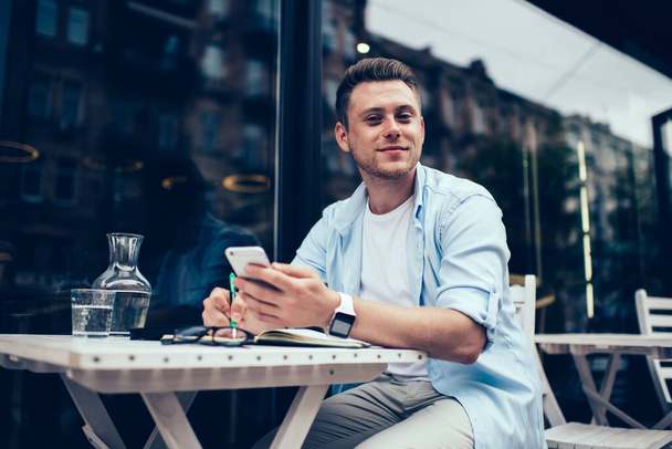 Handsome millennial man in casual outfit holding mobile phone while sitting at table and writing in notepad on street looking at camera - Foto, Bild