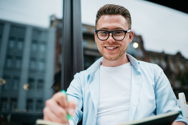 From below of smiling handsome millennial man in eyeglasses sitting at table on street terrace against window and looking at camera - Foto, afbeelding