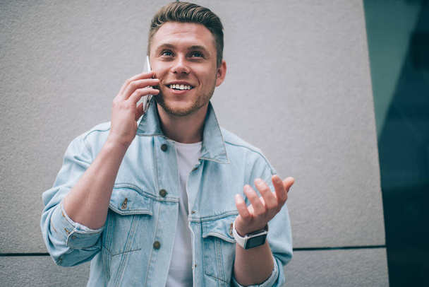 From below of trendy young man in denim jacket standing on street and smiling away while having call on mobile phone - Photo, Image