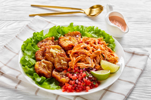 Spicy fried plantains with shredded chicken mixed with buffalo sauce, pico de gallo salsa, lime wedges on fresh lettuce leaves, on a white plate on a wooden background with cutlery, close-up - Foto, immagini