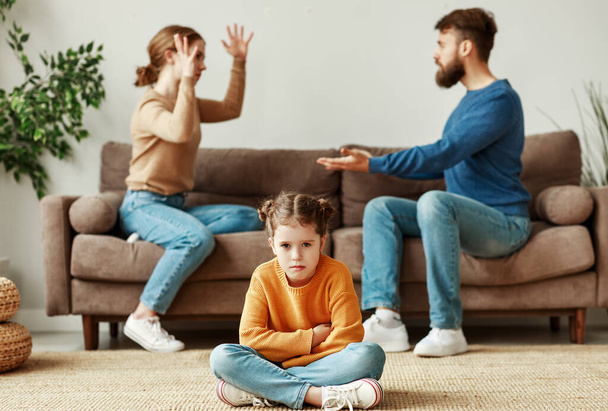 Mad parents sitting opposite each other on couch swearing loudly and gesturing with hands while upset little defenseless daughter sitting on floor and gloomy looking at camer - 写真・画像
