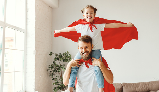 Sonriente barbudo joven sosteniendo linda chica en los hombros mientras juegan superhéroes juntos vestidos con capa de superhéroe rojo en roo ligero
 - Foto, Imagen