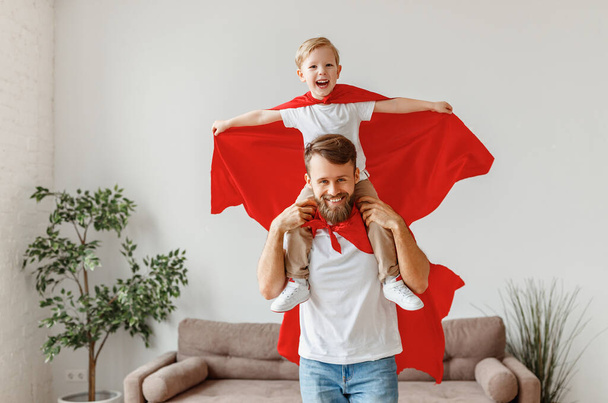 Cheerful bearded young father holding son on shoulders while having fun together dressed in red superhero cloak in living roo - Photo, Image