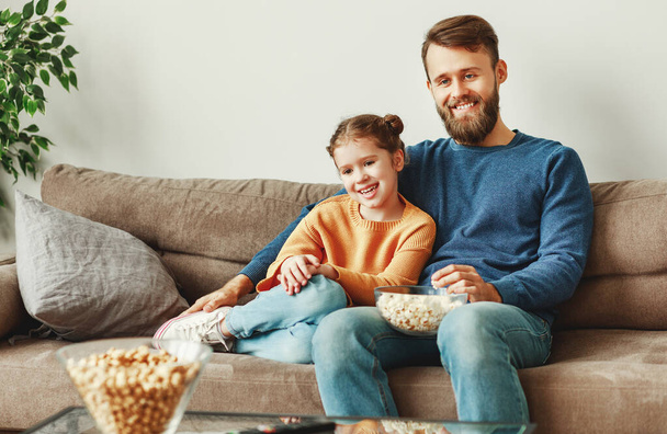 Smiling young dad with female kid sitting on couch against white wall in living room and cuddling while watching film during free tim - Photo, Image