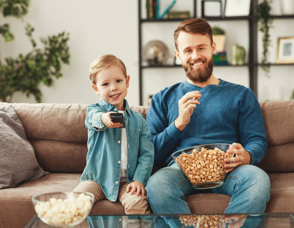 Ein bärtiger, fröhlicher junger Mann in lässiger Kleidung sitzt auf dem Sofa mit einem kleinen lächelnden Jungen, der eine lustige Show im Fernsehen anschaut und Popcorn isst, während Sohn den Sender wechselt - Foto, Bild