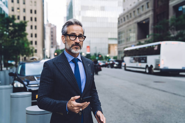 Attractive fashionable gray businessman with beard in stylish dark business suit standing near roadway with smartphone in hand and looking away in New York - 写真・画像