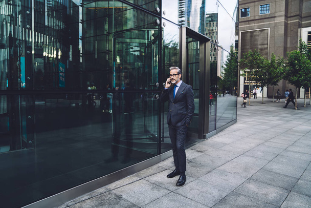 Middle aged businessman with beard in full growth in stylish dark business suit talking on phone near entrance to office building putting hand in pocket in New York - Φωτογραφία, εικόνα