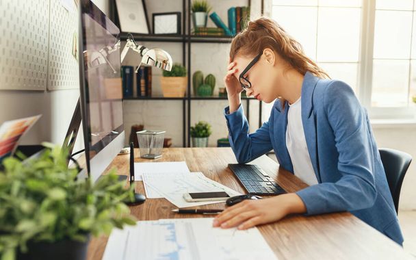 Frustrated female businesswoman in formal clothes with eyes closed trying to solve problem while sitting at table with notepad and computer and touching head in modern offic - Foto, immagini